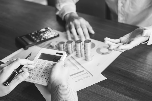 Hands using a calculator and pen over financial documents, with stacked coins and a person gesturing, illustrating cost management and financing options for plumbing and HVAC services.