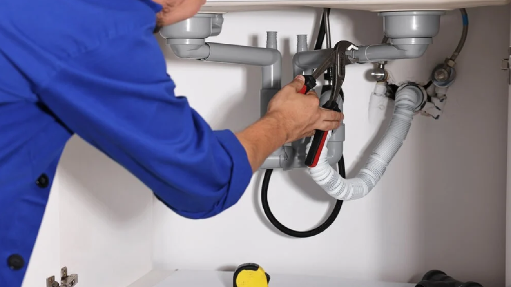 Plumber in blue shirt repairing kitchen sink plumbing under cabinet, showcasing professional service and quality workmanship by Durfee Plumbing & Heating LLC.
